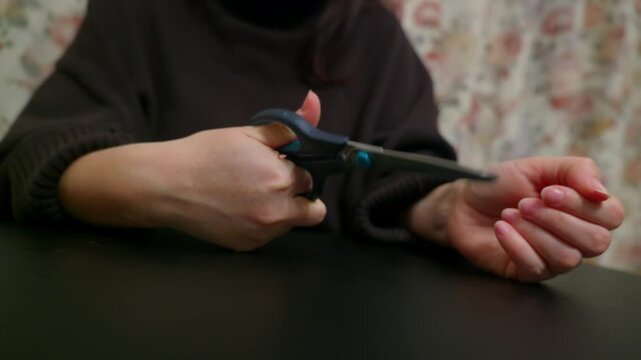 A woman in a dark sweater sits at a table, holding scissors to her wrist in a gesture symbolizing self harm, suicide, depression, and mental health crisis. Close up on hands