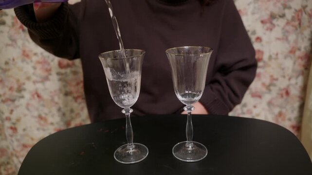 A person in a brown sweater pours clear water from a purple bottle into two elegant stemmed glasses with gold rims, sitting on a black table