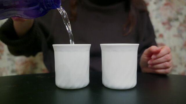 A closeup shot of a woman's hands pouring fresh, clear water from a blue reusable bottle into two white ceramic cups sitting on a black table. She fills one cup, then the other