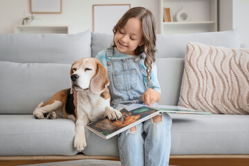 Naklejka premium Cute little girl with Beagle dog reading book on sofa in living room