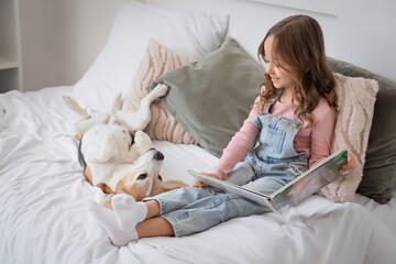 Naklejka premium Cute little girl with Beagle dog reading book on bed in bedroom