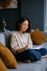 Obraz premium An Asian woman is sitting on a sofa, working with a notebook beside her.