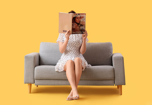 Beautiful young woman reading magazine on sofa against yellow background