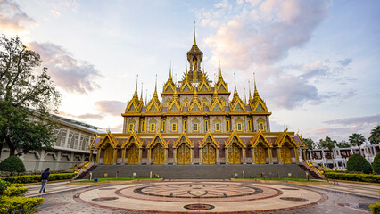 Naklejka premium An ornate Thai temple stands centered beneath a cloudy evening sky. Gold details and wide steps define the grand facade.
