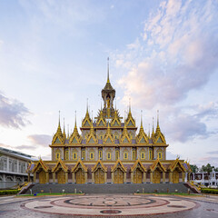 Naklejka premium An ornate Thai temple stands centered beneath a cloudy evening sky. Gold details and wide steps define the grand facade.
