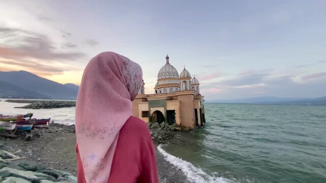 Muslim woman in a hijab stands by the shore, looking at the iconic damaged Floating Mosque of Palu, Central Sulawesi, a symbol of resilience after the 2018 tsunami.
