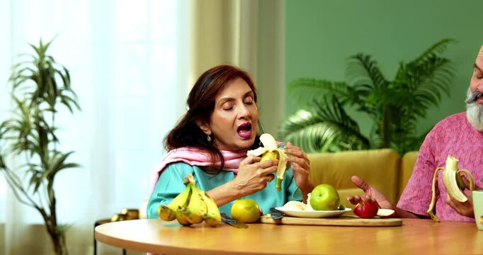 Senior Indian couple sitting at a breakfast table in their modern home while peeling and eating fresh yellow bananas together with smiles and casual conversation