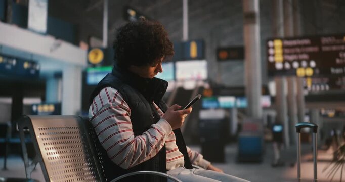 Sitting in Airport Lounge, Young Man With Curly Hair Uses Smartphone to Scan Digital Boarding Pass, Stays Focused on Screen, Scrolling Through Review Flight Details, Gate Number, and Departure Time.