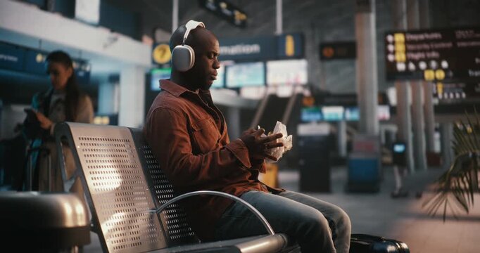 Black Man Sits Comfortably in Airport Waiting Area, Wearing Stylish Over Ear Silver Headphones. Guy Enjoying Hamburger, Taking Bites While Lost in Music. Concept of Lifestyle of Modern Passenger.