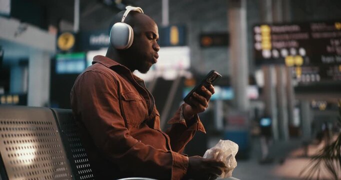 Black Man Sits Comfortably in Airport Waiting Area, Wearing Stylish Over Ear Silver Headphones. Guy Enjoying Hamburger, Taking Bites While Lost in Music. Concept of Lifestyle of Modern Passenger.