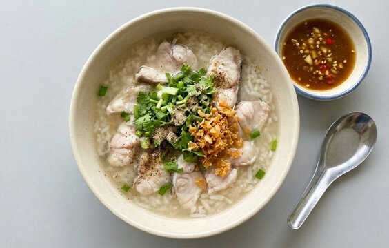 Authentic Thai fish rice soup topped with ginger, celery, and fried garlic. Served with spicy dipping sauce in a bowl over a clean grey background.
