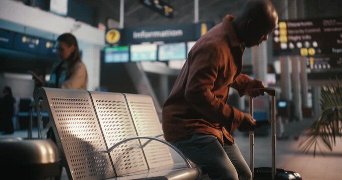 Black Male, Pulling Suitcase Enters Airport Waiting Area. Man Finds Seat and Uses Smartphone Scan Boarding Pass, Glances Between Device and Overhead Flight Information Boards Confirm at Correct Gate.