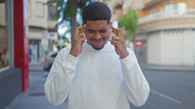 Man presses fingers to temples and squints with closed eyes on an urban street, white shirt visible; concentration focus.