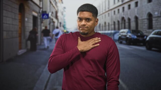 Hispanic man places hand on chest on a street wearing a maroon shirt and beard visible; sincerity humility.