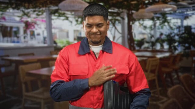 Man worker in red uniform holding toolbox places hand on chest inside building dining area with tables chairs plants and hanging lamps; pride duty.