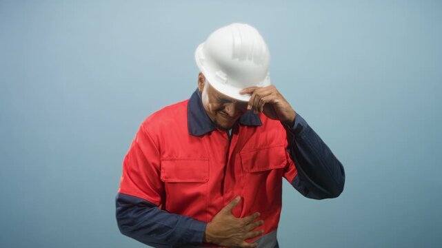 Hispanic construction worker in white hardhat and red uniform grips his stomach and tips his helmet with one hand in a studio; duty pain.