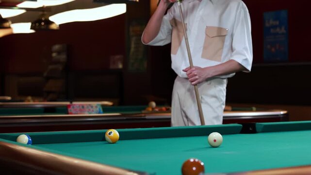 Young professional man billiard player rubs the end of the cue stick using special snooker chalk before billiards game important blow, standing in front of pool tables, sport activity at home indoors