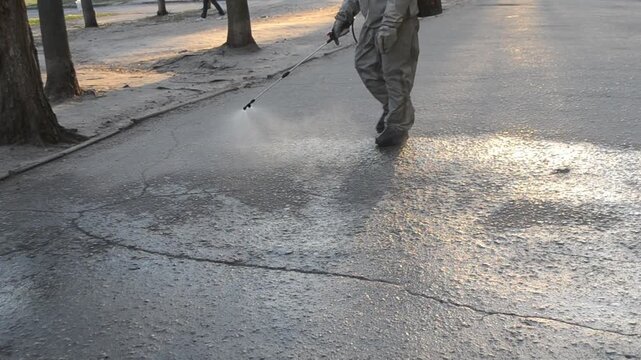 Man in airtight suit sprays disinfecting liquid chemicals antiseptic on street in city on day. Sanitation workers clean pavement, crosswalk. Sanitary measures in public place. Coronavirus pandemic