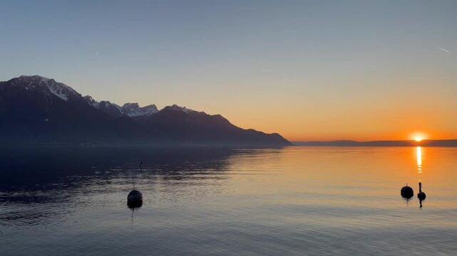 Peaceful sunset over Lake Geneva with calm water reflecting the warm orange sky and surrounding alpine mountains. Scenic tranquil landscape in Switzerland during golden hour, ideal for relaxation, tra