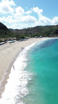Aerial view of the pristine Mawun Beach in Lombok, Indonesia, featuring turquoise crystal clear waters, white sandy coastline, and lush tropical hills under a bright sunny sky.