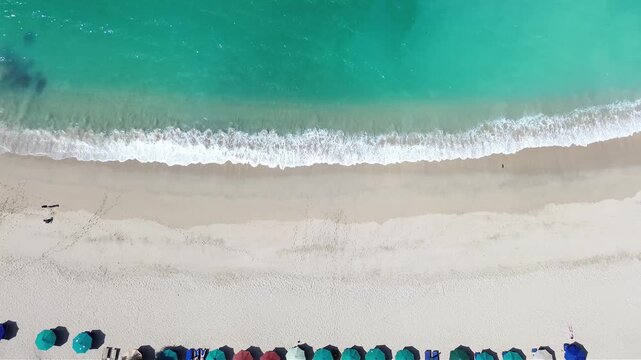 Aerial view of the pristine Mawun Beach in Lombok, Indonesia, featuring turquoise crystal clear waters, white sandy coastline, and lush tropical hills under a bright sunny sky.