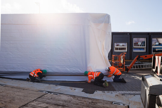 Three construction workers in full safety gear collaborating to wrap a prefabricated office unit, captured with sunlight flaring into the lens.