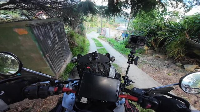 Relaxed first-person perspective from a motorcycle's handlebars cruising down a paved road in a lush, green residential mountain area.