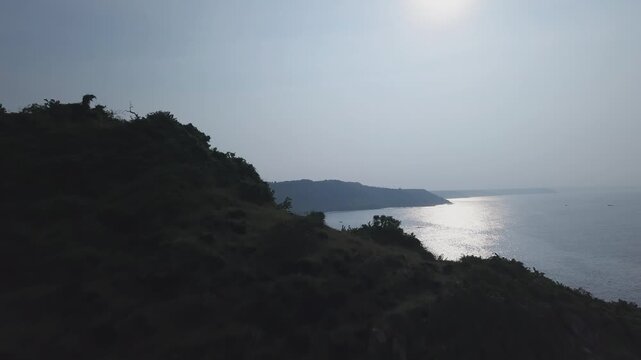 Drone moves forward across forested hillside near Nivti Fort to reveal the Malvan coastline, Nivti Beach, and the Arabian Sea under hazy daylight skies in Maharashtra, India.