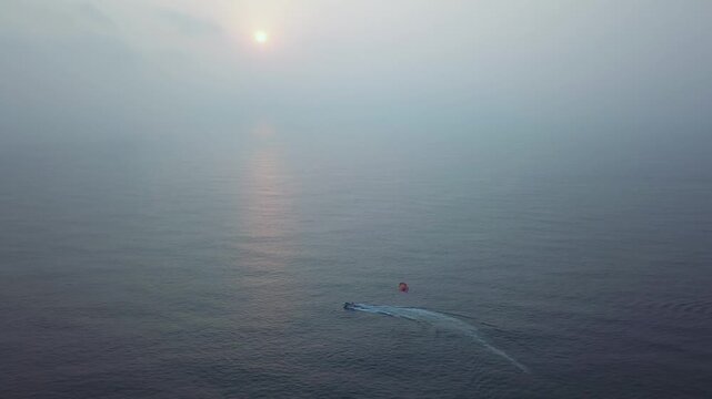 Parasailer towed by speedboat across calm ocean waters during sunset at Candolim Beach in Goa, India. Hazy golden light reflects on water surface as adventure water sport activity occurs offshore.