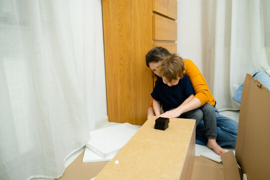 A mom is assembling a shelving unit in the room, with her toddler sitting comfortably on her lap. A scene of modern parenthood combining care and home improvement tasks.
