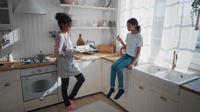 Cheerful black woman holding hot baking sheet with cookies, talking with girl. Mother and daughter cooking sweets at home, enjoy delicious hypoallergenic homemade bakery, traditional healthy food