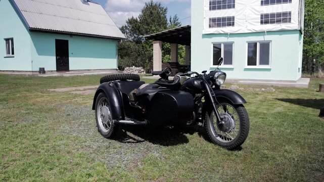 Classic black vehicle, motorcycle with sidecar, stands stationary on well-maintained lawn. Background features two light blue houses, one with white facade covering upper floor windows.