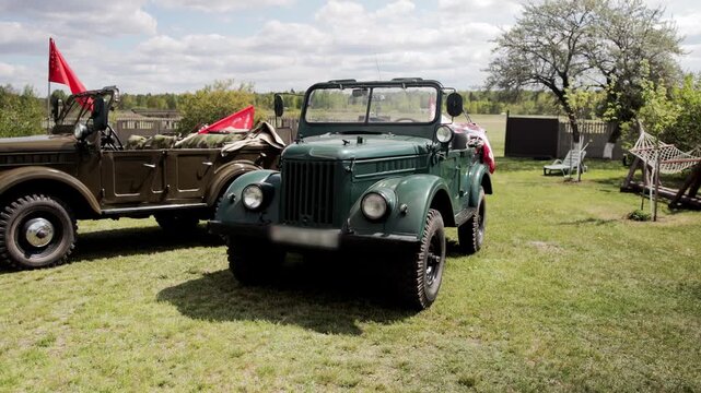 Two vintage dark green military vehicles on grass field under partly cloudy sky. Open-top utility transport with red flags, outdoor setting for historical display, evocative of May 9th victory day com