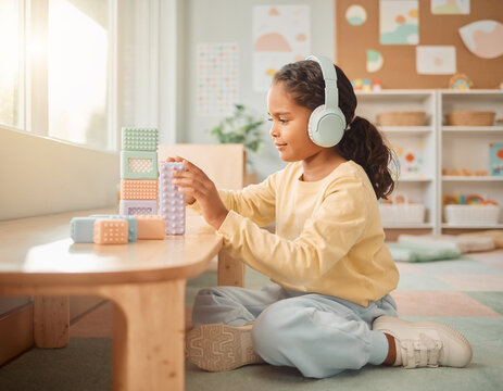 Young girl wearing noise-canceling headphones calmly playing with textured pastel sensory blocks at a low wooden table in a bright sunlit therapy room
