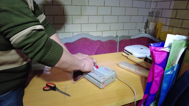 A person is using a vacuum sealer to pack food in bags on a table. The setting is a simple home kitchen with various items around. The task involves preparing food for storage.