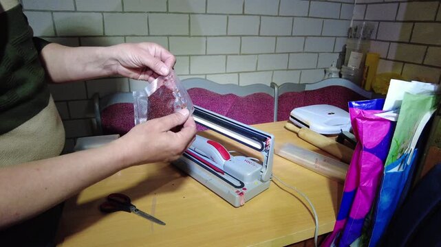 A person demonstrates how to use a food sealer on a wooden kitchen table. They prepare items for sealing while tools and bags are nearby.