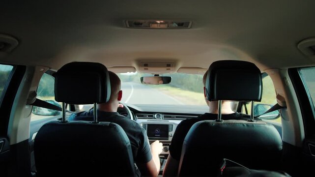 Passenger perspective of two men driving modern car on open road, vehicle interior visible with dashboard display and steering wheel, green trees lining road, daytime travel