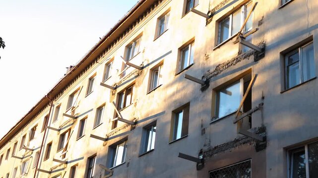 Exterior view of multi-story apartment building undergoing renovation, showing remnants of demolished balconies. Scaffolding elements protrude from weathered facade, bathed in warm sunlight.