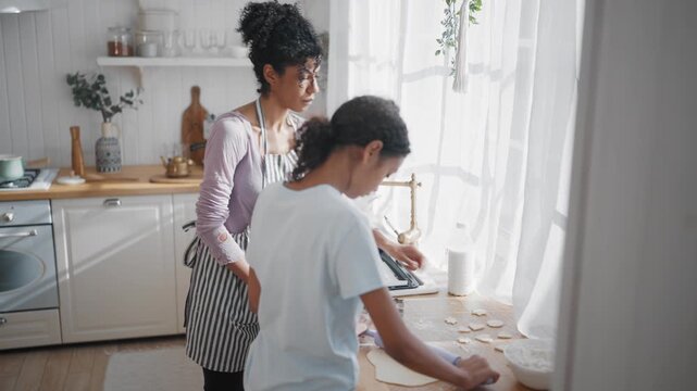 Cute african american teen girl cooking cookies, mother helping, childhood. Careful adult woman teaching her talented little daughter to cook, baking tasty ginger cookies for christmas or easter fest