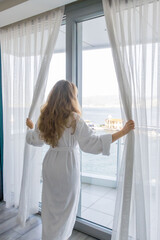 Obraz premium Woman opening curtains in apartment near large window with sea view. Morning routine, home lifestyle, natural light, daily life moment indoors