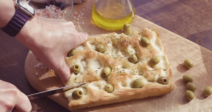Anonymous person cutting fresh homemade olive focaccia on wooden board with copy space. High-angle video captures hands slicing traditional Italian flatbread with knife on rustic table. Footage