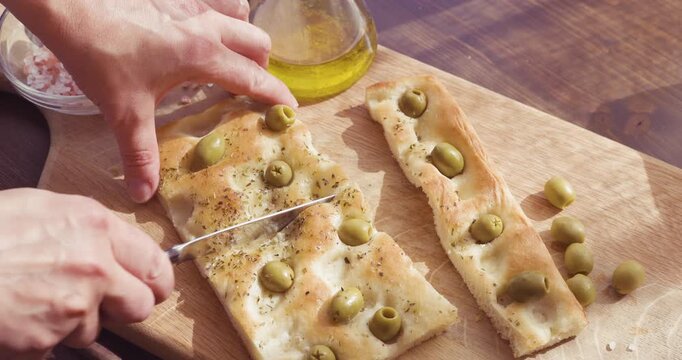Anonymous person cutting fresh homemade olive focaccia on wooden board with copy space. High-angle video captures hands slicing traditional Italian flatbread with knife on rustic table. Footage