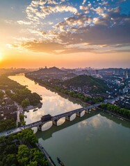 Fototapeta premium A stunning aerial view captures a historic bridge spanning a tranquil river at sunset, framed by a vibrant cityscape and lush greenery.