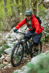 Fototapeta premium Man mountain biking on a rocky forest trail in red jacket with helmet and backpack
