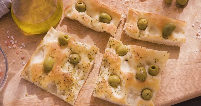 Traditional Italian olive focaccia slices on wooden board with pink salt and olive oil. High-angle video captures authentic Mediterranean bakery texture and rustic homemade aesthetic under warm