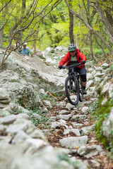 Naklejka premium Mountain biker riding a rocky forest trail wearing a red jacket and helmet jumping over stones