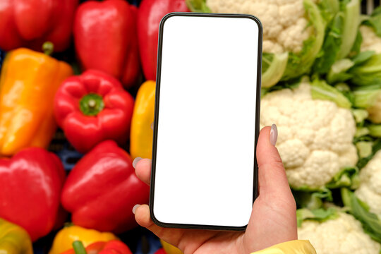 Hand holds a smartphone with a blank white screen above fresh bell peppers and cauliflower at a grocery market, suggesting mobile grocery shopping, recipe planning, produce comparison and healthy food
