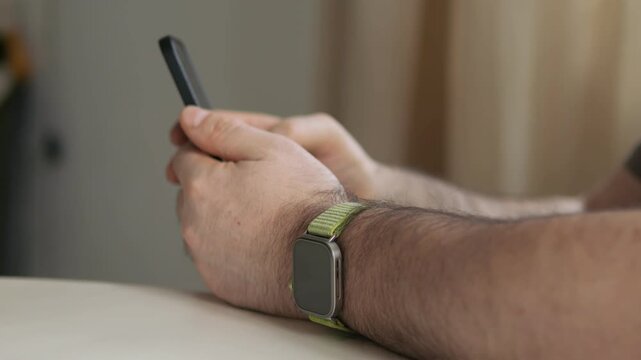 Man holds smartphone with both hands. Wears green watch strap. Rests arms on table. Background softly blurred. Indoor setting with neutral tones