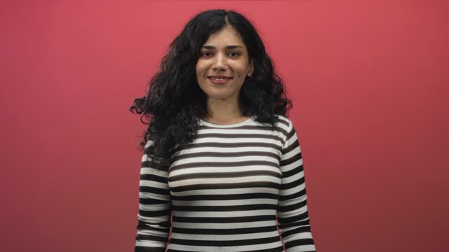 Young arab woman wearing a striped top smiles with eyes closed showing cheek piercings and bare face in a pink studio setting; contentment.