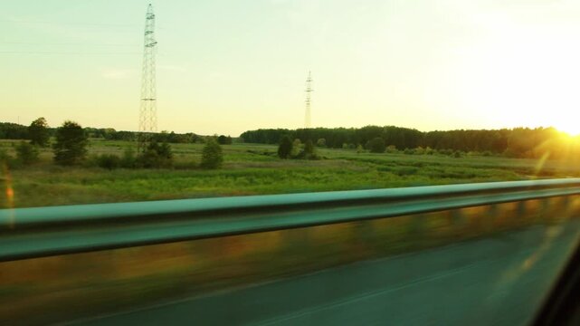 Scenic car journey perspective of sun setting over vast green meadow and dark forest, blur of speed visible on metal guardrail, power transmission towers stand tall in open country.